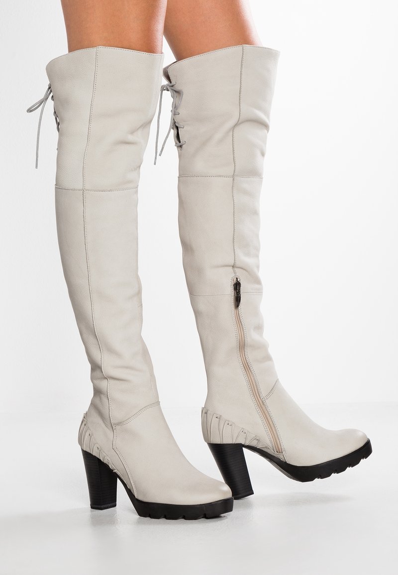 Białe kozaki za kolano ze skóry o fakturowanej powierzchni, bocznym zamkiem błyskawicznym i czarnymi masywnymi obcasami. Z tyłu ozdobione sznurowaniem.