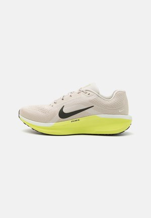 Nike hardloopschoen in lichtbeige mesh met zwarte swoosh, gele tussenzool en gestructureerde zool. Ontworpen voor comfort en ondersteuning.