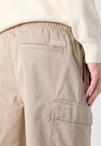 Calções cargo bege feitos de material leve, com uma cintura elástica, bolsos laterais e uma pequena etiqueta com o logótipo vermelho da Calvin Klein.