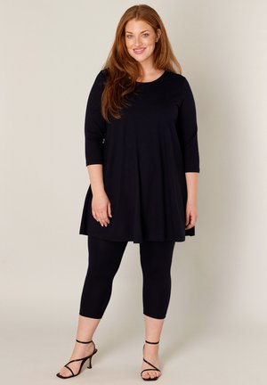 Vrouw met rood haar, gekleed in een zwarte tuniek met lange mouwen, zwarte cropped leggings en zwarte sandalen met hoge hakken en bandjes, staand en glimlachend.