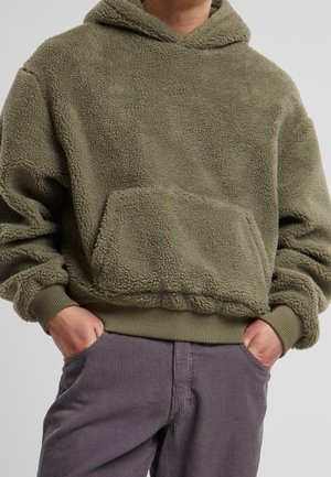 Personne portant un sweat à capuche en polaire vert olive avec une poche avant et un pantalon en velours gris, mains dans les poches.