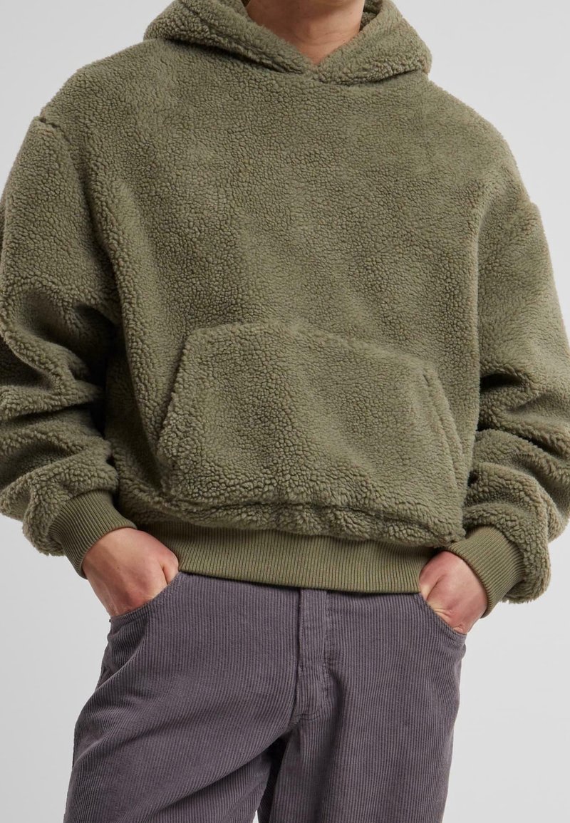 Personne portant un sweat à capuche en polaire vert olive avec une poche avant et un pantalon en velours gris, mains dans les poches.