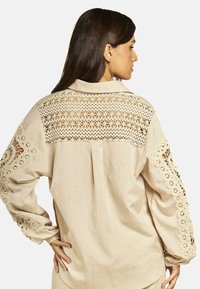Camisa beige de botones con inserciones de encaje en las mangas y la espalda. Presenta un corte relajado y mangas largas con un detallado borde de encaje.