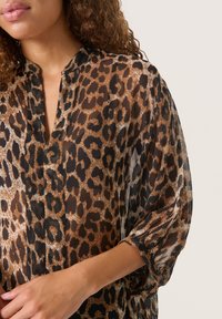 Bluse med leopardprint, v-hals, gennemsigtigt stof, løse ærmer og en blanding af brune, sorte og crème farver. Subtile teksturdetaljer er til stede.