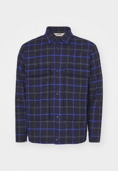 Ternet jakke lavet af tekstureret stof, med en mørk navy baggrund og blå samt grå firkanter, to brystlommer og trykknapper.