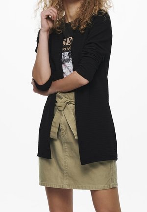 Femme aux cheveux bouclés porte un cardigan noir texturé, un t-shirt graphique et une jupe beige avec ceinture nouée, posant les bras croisés.
