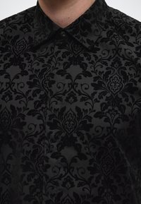 Must särk, millel on keerukas sametdamaskiki muster. Sellel on terav krae ja nööpidega esipaneel, sile tekstuur ning luksuslik välimus.