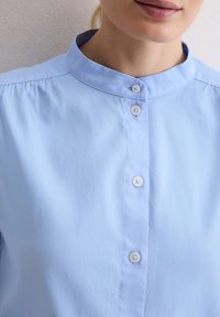 Kobieta w jasnoniebieskiej koszuli z kołnierzykiem i białymi guzikami, widoczna od szyi do klatki piersiowej na jednolitym tle.