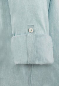 Puño de camisa de lino azul claro con un solo botón blanco. Textura visible de la tela, que presenta un acabado suave y ligeramente arrugado.