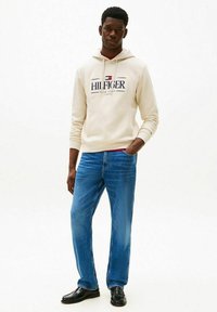 Felpa con cappuccio color beige chiaro con logo, jeans blu e scarpe nere. La felpa ha un cordino e polsini a coste; i jeans sono a taglio dritto.