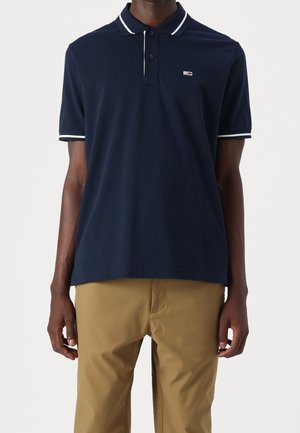 Homme portant un polo bleu marine à manches courtes avec une bordure blanche et un petit logo, associé à un pantalon beige, debout sur un fond uni.
