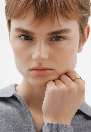 Anillo de oro con acentos circulares que se lleva en una mano apoyada sobre un cuello de punto gris. El primer plano muestra una textura de piel suave y ojos azules visibles.