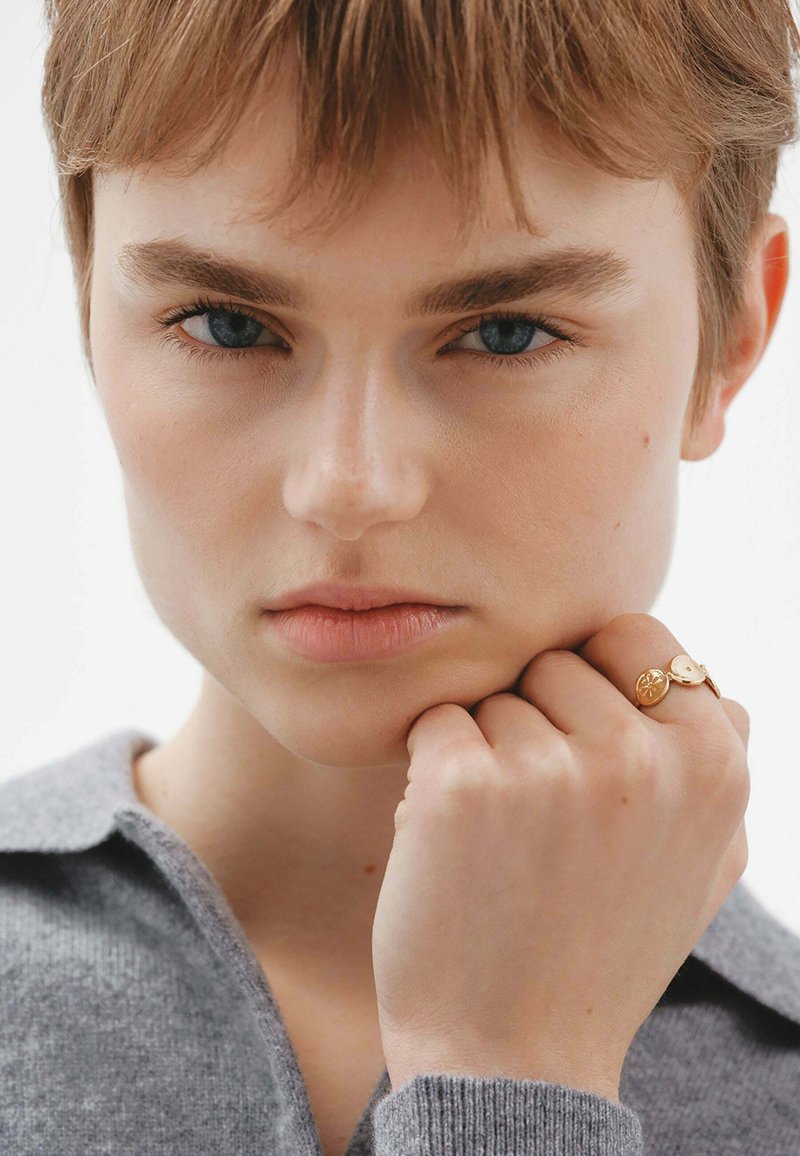 Anillo de oro con acentos circulares que se lleva en una mano apoyada sobre un cuello de punto gris. El primer plano muestra una textura de piel suave y ojos azules visibles.