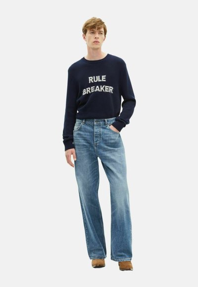 Maglione navy con testo "RULE BREAKER", abbinato a jeans a gamba larga blu chiaro e scarpe marroni. Pose in piedi inclinata con le mani in tasca.