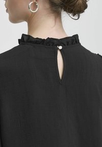 Top nero realizzato in tessuto testurizzato, con scollatura arricciata e chiusura a chiave sulla schiena decorata con un piccolo bottone.