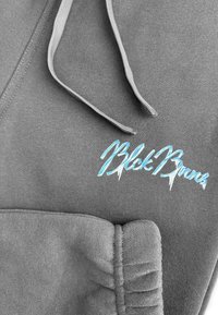 Grijze hoodie stof met een zachte textuur. Bevat een turquoise geborduurd logo met de tekst "Blok Buns." De koorden zijn grijs.