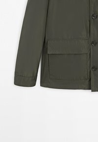 Manica di giacca verde scuro e parte anteriore inferiore con grande tasca a patta e bottoni neri su sfondo bianco.