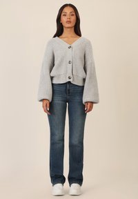 Cardigan grigio lavorato a maglia con scollo a V, chiusura con bottoni e maniche ampie, abbinato a jeans blu e sneakers bianche.