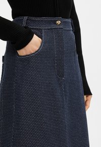 Σαλοπέτα τζιν σε σκούρο μπλε με υφασμάτινο σχέδιο, διαθέτει πλαϊνές τσέπες και χρυσή κουμπί αξεσουάρ στη μέση.