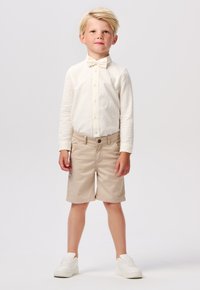 Chemise à manches longues claire, shorts beiges et baskets blanches. Noeud papillon en détail d'accent. Tissu lisse avec une coupe décontractée.