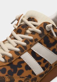 Κοντινό πλάνο αθλητικού παπουτσιού με μοτίβο λεοπάρδαλης, λευκά κορδόνια και δύο λευκές ρίγες στο πλάι, με επενδεδυμένη γλώσσα και καφέ σόλα.