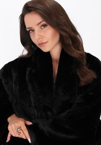 Giovane donna con capelli castani ricci che indossa un cappotto di pelliccia sintetica nera, mentre tiene il colletto con la mano sinistra decorata con un anello d'argento di grande impatto.