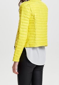 Yellow quilted jacket with vertical stitch pattern, cropped design, front zip, and long sleeves. Worn over a white top and black pants.