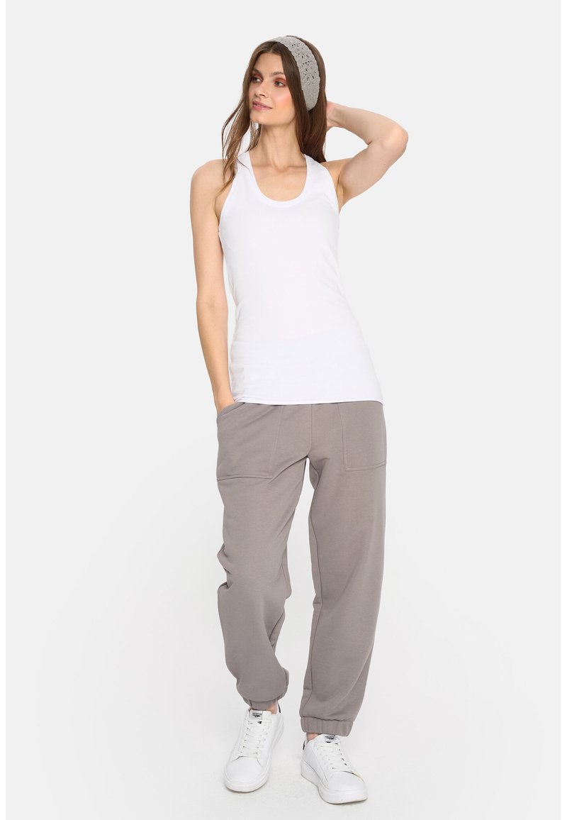 Biały top na ramiączkach z dekoltem typu scoop, w połączeniu z jasnoszarymi spodniami dresowymi z dwoma przednimi kieszeniami. Modelka ma na sobie białe trampki.