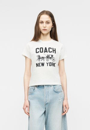 Młoda kobieta w białej koszulce Coach New York z grafiką konia i powozu oraz jasnoniebieskich, wysokich jeansach, stojąca na jednolitym tle.