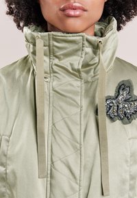 Giacca trapuntata verde chiaro con colletto alto, coulisse e una toppa decorativa ricamata con dettagli argentati e neri.