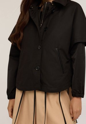 Femme portant une veste noire boutonnée avec des détails de fermeture éclair sur une jupe beige à rayures verticales noires.