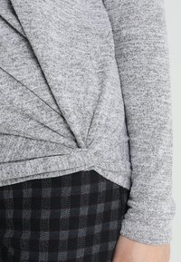 Maglione grigio a maglia con design attorcigliato in vita, texture liscia e maniche lunghe, abbinato a pantaloni a quadri neri.