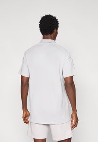 Ανοιχτό γκρι πόλο μπλουζάκι με κοντά μανίκια και γιακά, με ομαλή υφή και ίσια ραφή στο κάτω μέρος. Η πίσω όψη παρουσιάζει ελάχιστες λεπτομέρειες.