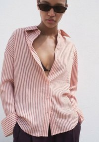 Chemise boutonnée rose clair avec de fines rayures verticales rouges, un fit décontracté et des poignets retroussés. Assortie à des lunettes de soleil noires.