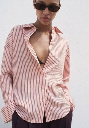 Helles rosa Button-up-Hemd mit dünnen vertikalen roten Streifen, lockerer Passform und aufgerollten Ärmeln. Kombiniert mit einer schwarzen Sonnenbrille.