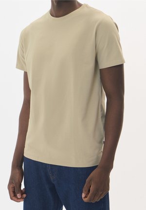 Homme portant un t-shirt beige uni à manches courtes et un jean bleu foncé, debout devant un fond clair uni.