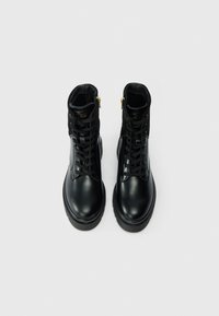 O pereche de boots din piele neagră până la gleznă, cu șireturi și fermoare pe lateral, expuse din față pe un fundal alb.