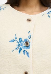 Kremasta tkanina z modrim cvetličnim vezenjem in tremi okroglimi bež gumbi na zgornjem delu prsnega koša in območju izreza ženske.