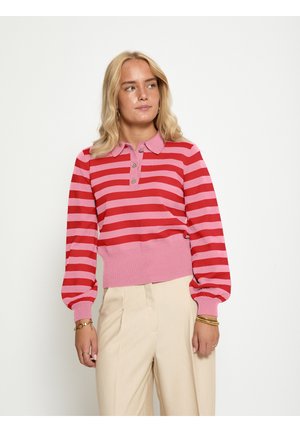 Kvinde med blondt hår iført en lyserød og rødstribet langærmet polotrøje og beige højttaljede bukser, stående mod hvid baggrund.