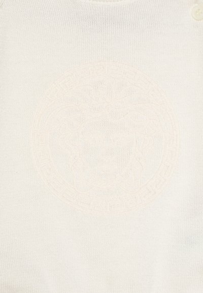 Balta adīta apģērba daļa ar reljefu apļa dizainu, kurā centrā ir sejas motīvs, ko ieskauj ģeometriskais raksts.