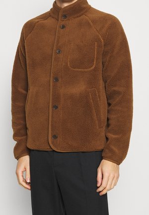 Chaqueta de forro polar marrón con cuello, botones frontales y un bolsillo en el pecho. Presenta una superficie texturizada y mangas raglán.