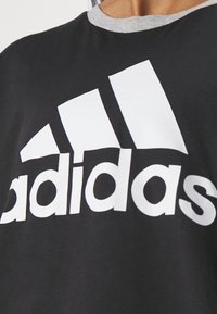 Černé bavlněné tričko s kulatým šedým výstřihem, s výrazným bílým logem Adidas a třemi pruhy v trojúhelníkovém designu.