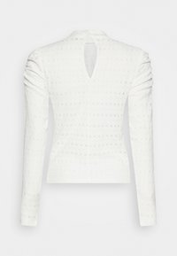 Blusa bianca a maniche lunghe con motivo ad occhio di pernice, maniche arricciate e una piccola apertura a goccia nella parte superiore della schiena.