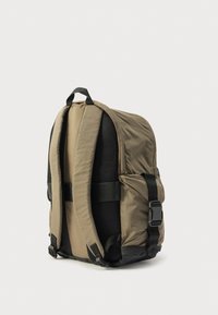 Khaki színű hátizsák sima szövetborítással, hálós párnázattal, állítható pántokkal és fekete díszítésekkel. Felül fogantyúval és oldalsó zsebekkel rendelkezik.