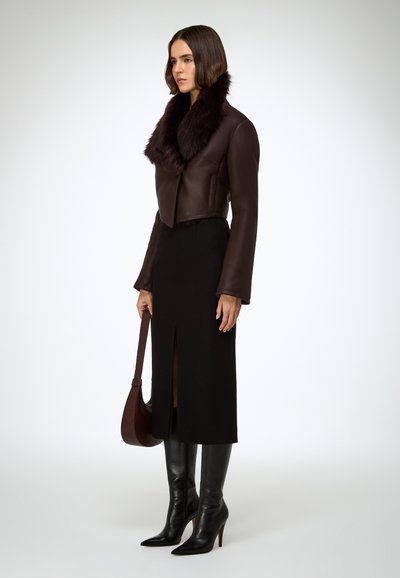 Giacca corta in pelle marrone con colletto in pelliccia, abbinata a una gonna nera con spacco e stivali neri con tacco a spillo al ginocchio. Borsa marrone.