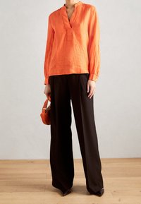 Blouse en lin orange avec un col en V et des manches longues, associée à un pantalon noir à jambes larges. Le modèle tient un petit sac à main orange.