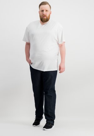 White short-sleeve V-neck t-shirt, dark blue jeans, and black sneakers. Model stands facing forward on a plain white background.