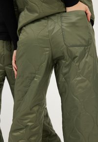 Pantalon matelassé vert olive avec une texture lisse et brillante et un motif de couture ondulé. Il possède une poche arrière et un design à coupe décontractée.
