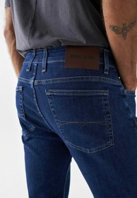 Jeans de mezclilla azul oscuro con una etiqueta de cuero marrón que dice "SALSA JEANS". Presenta dos bolsillos traseros con detalles de costura.
