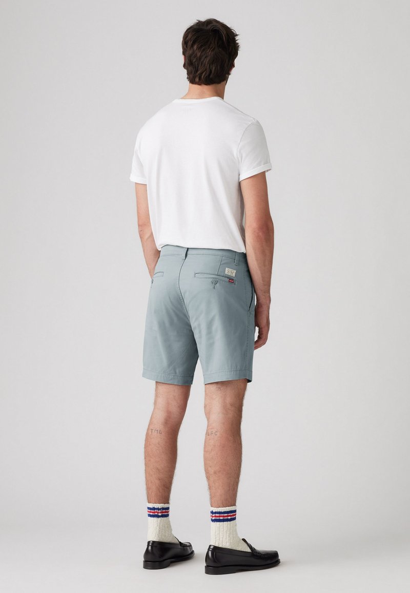 Shorts bleu clair avec poches latérales, portés avec un t-shirt blanc, des chaussettes crème à rayures rouges et bleues, et des mocassins noirs.